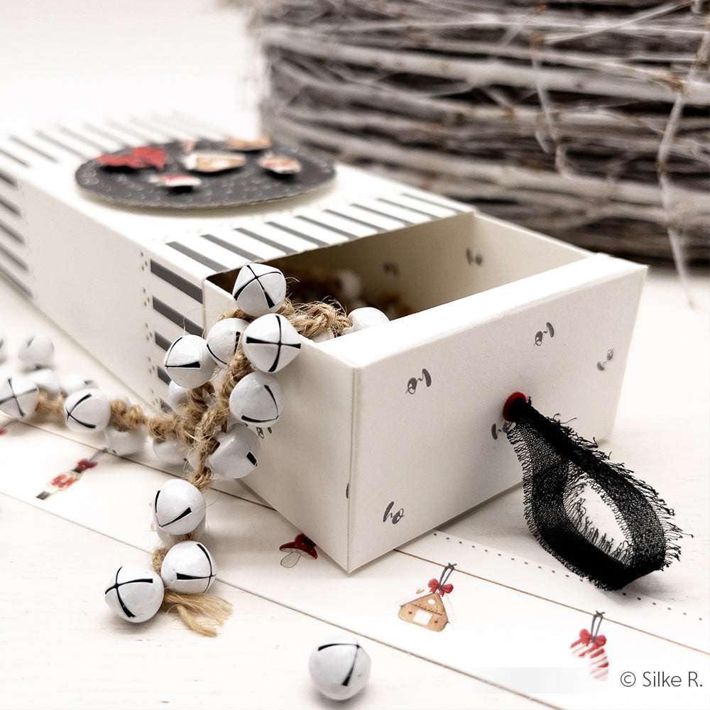 Eine gestreifte Streichholzschachtel von Alexandra Renke mit Jingle Bells an Schnüren, schwarzem Band und festlichen Akzenten liegt auf einer weißen Fläche - ideal für Papierprojekte oder in Kombination mit Grußkarten für einen charmanten Weihnachtslook.