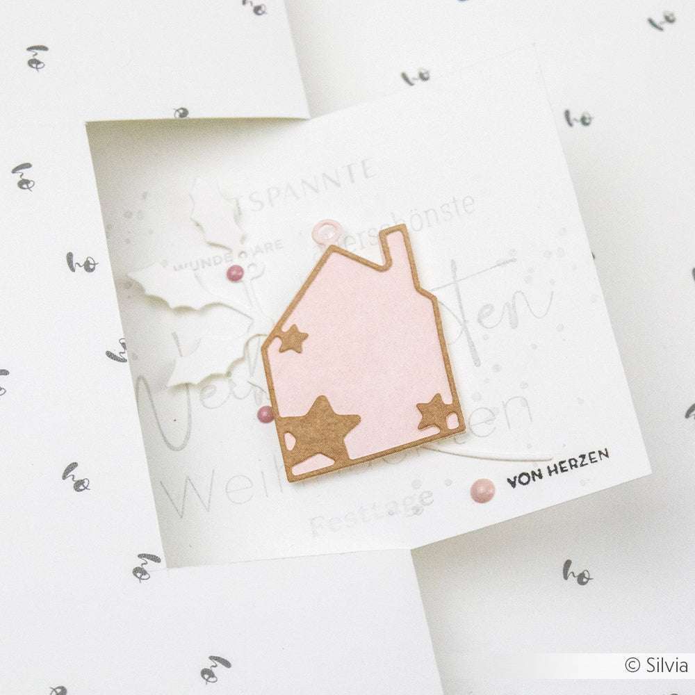 Ein rosafarbenes Papierhaus mit Sternausschnitten auf einer weißen Karte von Alexandra Renke, verziert mit Blättern und rosafarbenen Punkten - ideal für kreative Papierprojekte mit Designerpapier.