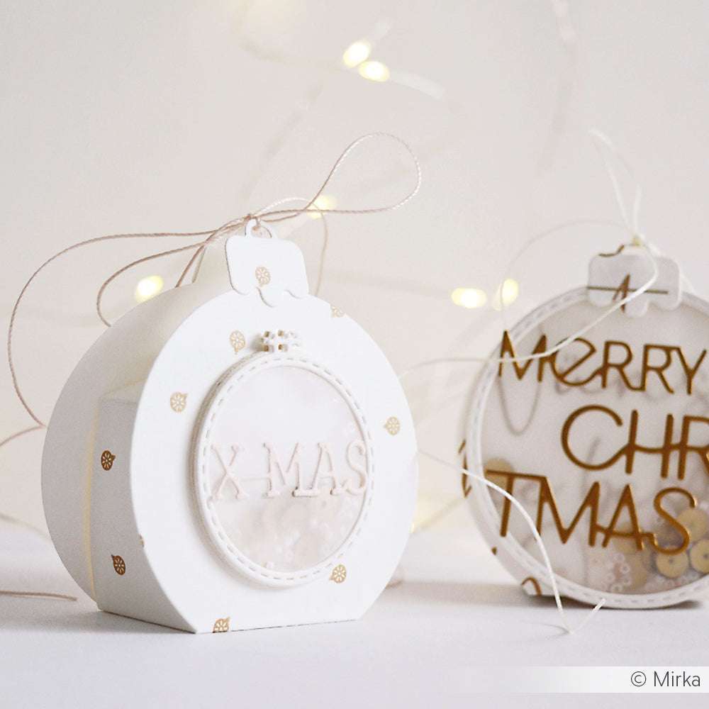 Zwei weiße Weihnachtsornamente mit goldenen Akzenten und den Texten "XMAS" und "MERRY CHR XMAS" von Alexandra Renke sorgen für einen eleganten Touch - perfekt für Ihre Papierprojekte oder in Verbindung mit festlichem Designpapier. Dahinter leuchtet eine weiche Lichterkette.