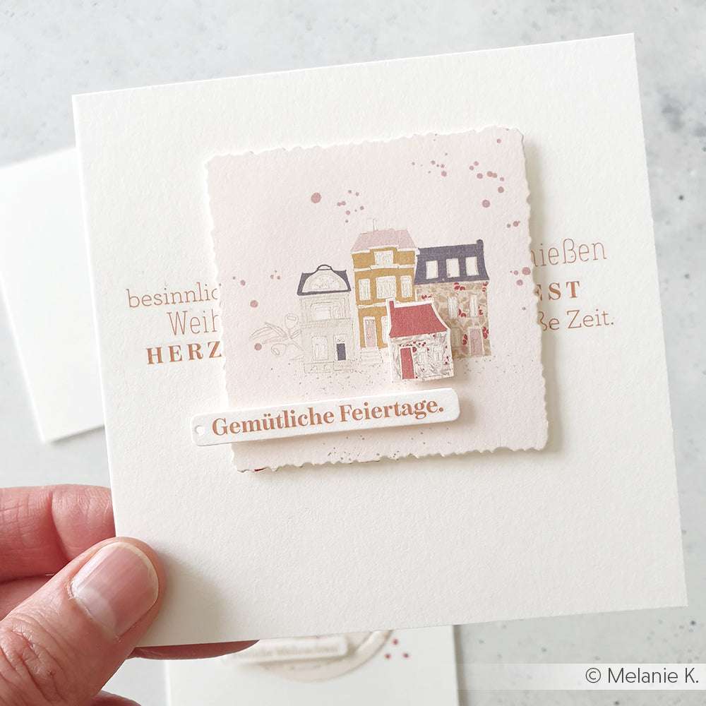 Eine Hand hält eine Feiertagskarte von Alexandra Renke mit gemusterten Häusern und dem deutschen Text "Gemütliche Feiertage" - eine perfekte Inspiration für Papierprojekte oder Ihr Lieblingsdesignpapier.