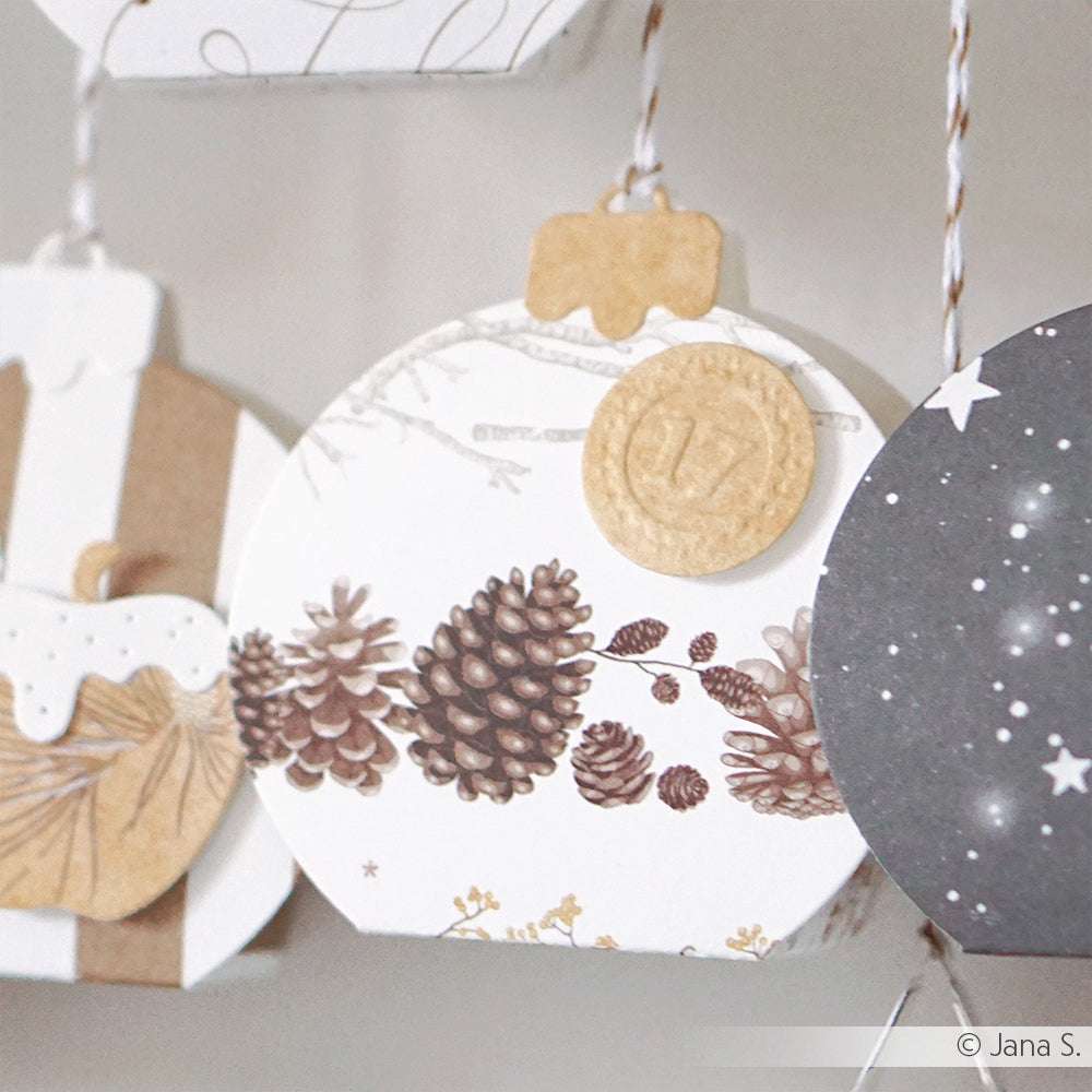 Rundes Ornament mit Tannenzapfenmotiv auf Designpapier von Alexandra Renke, Goldmünze mit der Nummer 17, an einer Schnur hängend, neben anderen Papierprojekten und Ornamenten.