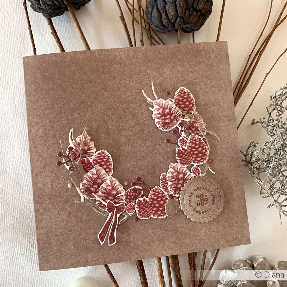 Quadratische braune Karte von Alexandra Renke, hergestellt mit Designpapier und verziert mit einem Kranz aus Tannenzapfen und Beeren sowie einem runden "Ho Ho Ho Frohe Weihnachten"-Anhänger - ideal für festliche Papierprojekte.