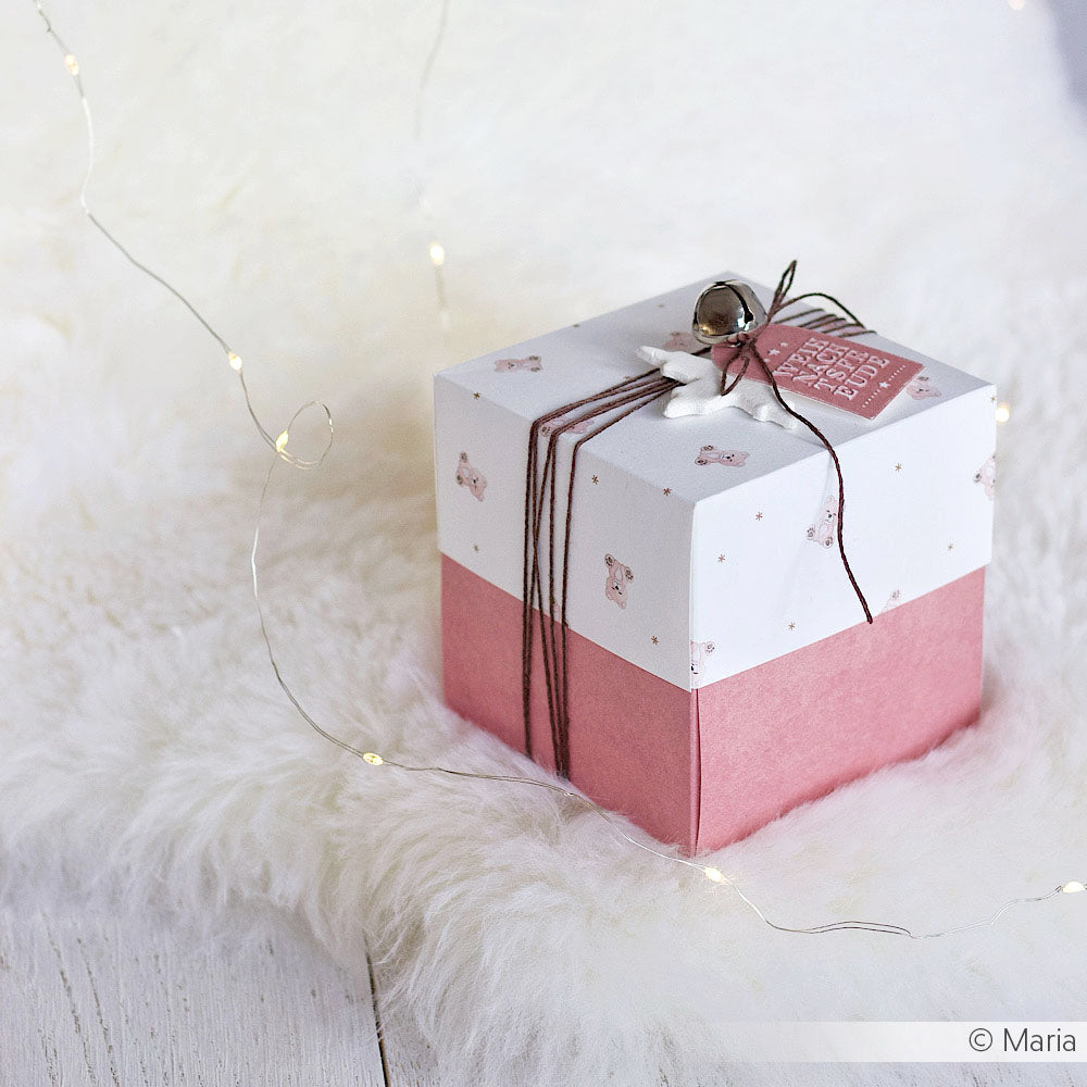 Eine rosa-weiße Alexandra Renke Geschenkbox mit Schnur und Glöckchen sitzt auf einer flauschigen weißen Fläche mit Lichterkette, ideal für Papierprojekte oder zum Kombinieren mit Grußkarten aus dem Designpapier Teddybär-Gewusel Rosa.