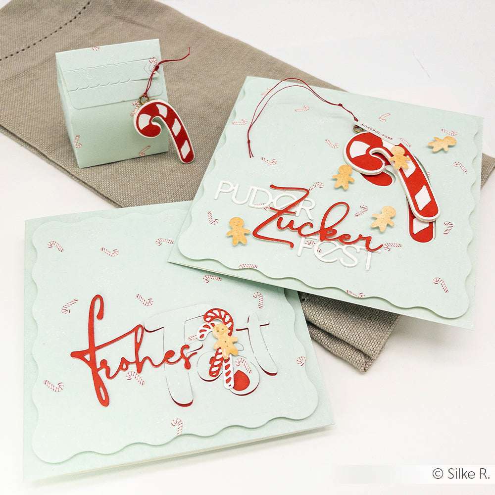 Grußkarten und eine Geschenkschachtel mit Zuckerstangen- und Lebkuchenmotiven, die mit einem festlichen deutschen Text versehen sind, verwenden Designpapier aus der Kollektion Zuckerstangen-Gewusel Mint von Alexandra Renke - ideal für Papierprojekte auf einem neutralen Stoffhintergrund.
