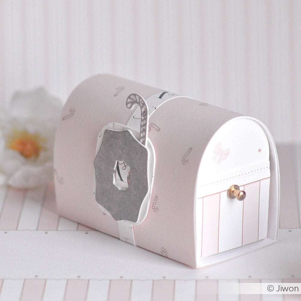 Eine pastellrosa-weiße Dekobox mit Metallverschluss aus Alexandra Renke Designpapier ruht auf einer weichen, gestreiften Oberfläche.