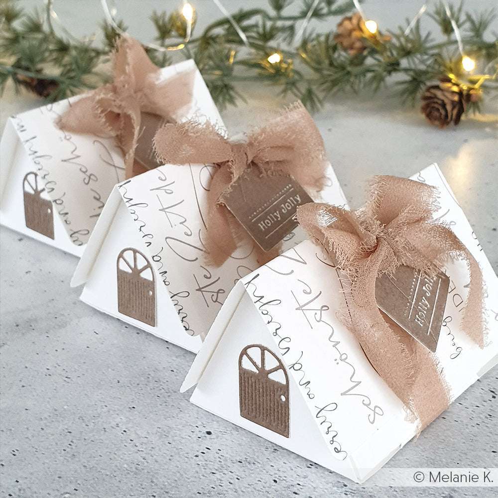 Drei weiße Geschenkboxen in Hausform von Alexandra Renke, umwickelt mit braunen Schleifenbändern und Holly Jolly-Anhängern, stehen vor Lichterketten - eine ideale Ergänzung zu Grußkarten oder Designpapier für festliche Geschenke.