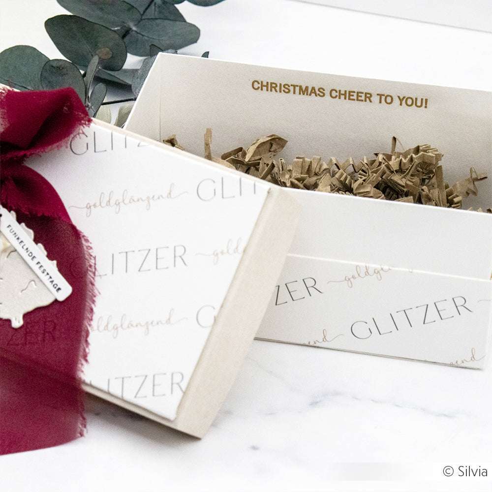 Alexandra Renke Geschenkkarton mit burgunderrotem Band, geöffnet, um die Füllung aus geschredderten Papierarbeiten und die festliche Botschaft zu zeigen: WEIHNACHTSFREUDE FÜR SIE!.