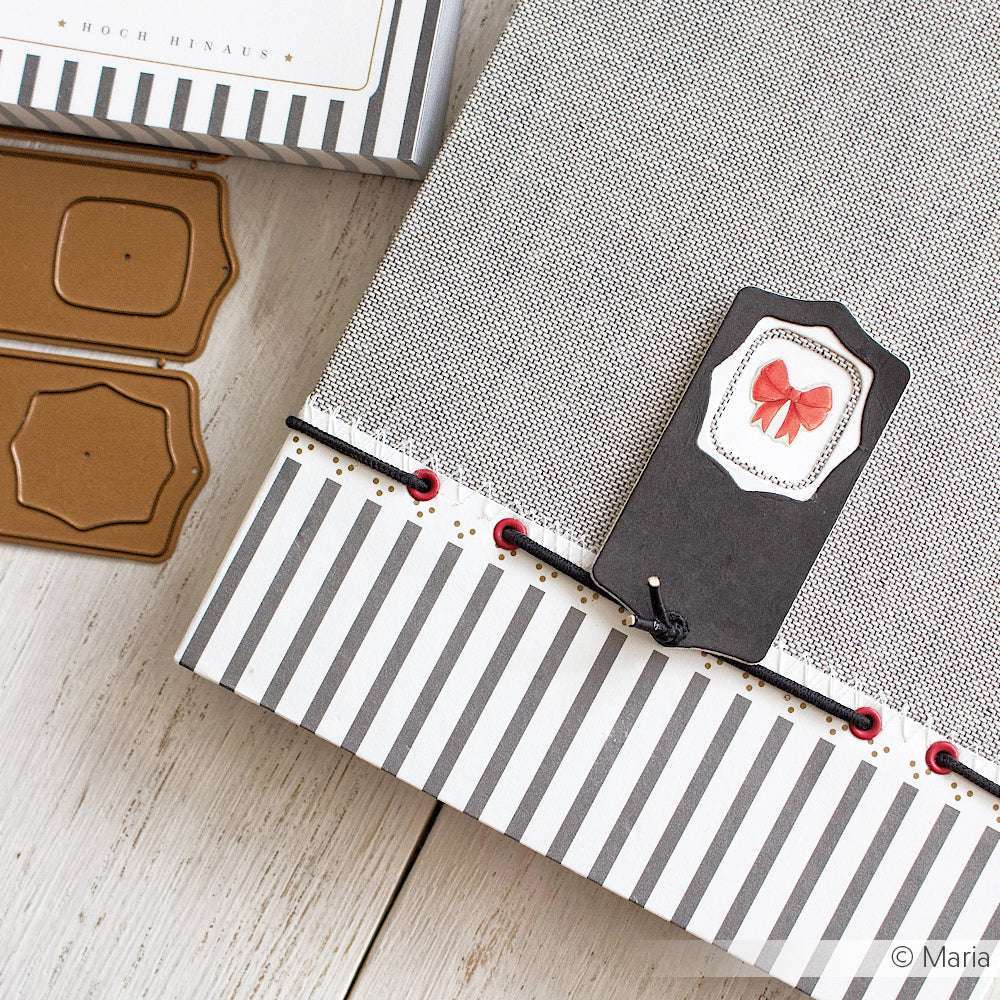 Gestreiftes Scrapbook von Alexandra Renke mit Buchbinden-Details und einem schwarzen Anhänger mit rosa Schleife, daneben braune Etikettenausschnitte und Minialben-Ideen, ausgestellt auf einer Holzfläche.