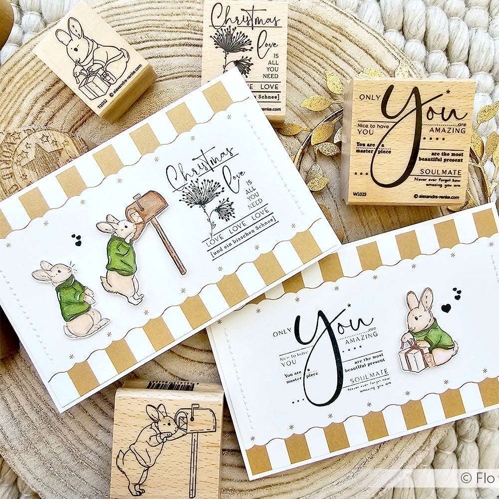 Zwei handgefertigte Alexandra Renke Karten mit niedlichen Hasen, weihnachtlichem Text, Stern-Bordüre Gold und Holzstempeln liegen auf einer gewebten Oberfläche - perfekt für Ihre Papierprojekte.