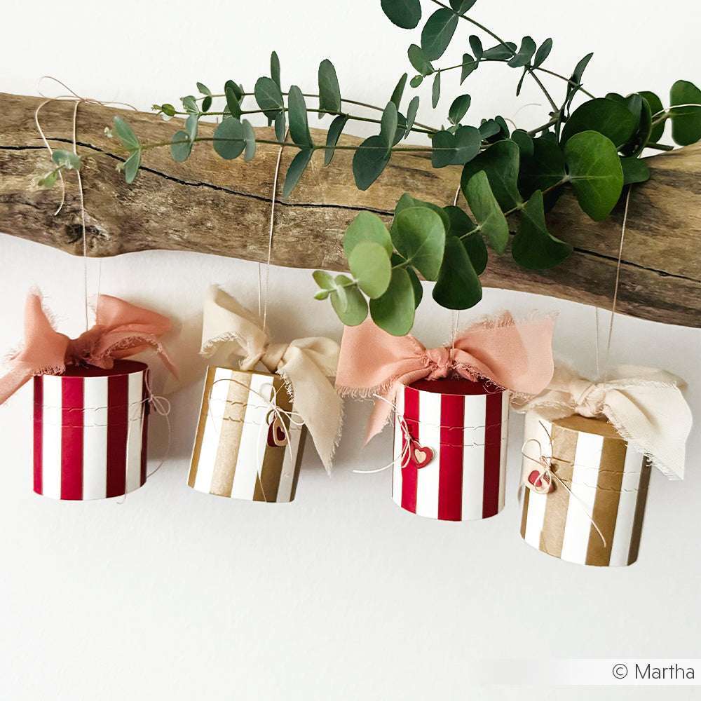 Vier gestreifte Ornamente mit Schleifen - zwei rot-weiße, zwei gold-weiße - hängen an einem grünen Zweig. Die mit Alexandra Renke Designpapier gefertigten und mit Stern-Bordüre Gold akzentuierten Papierprojekte bringen festlichen Charme in jede Einrichtung.