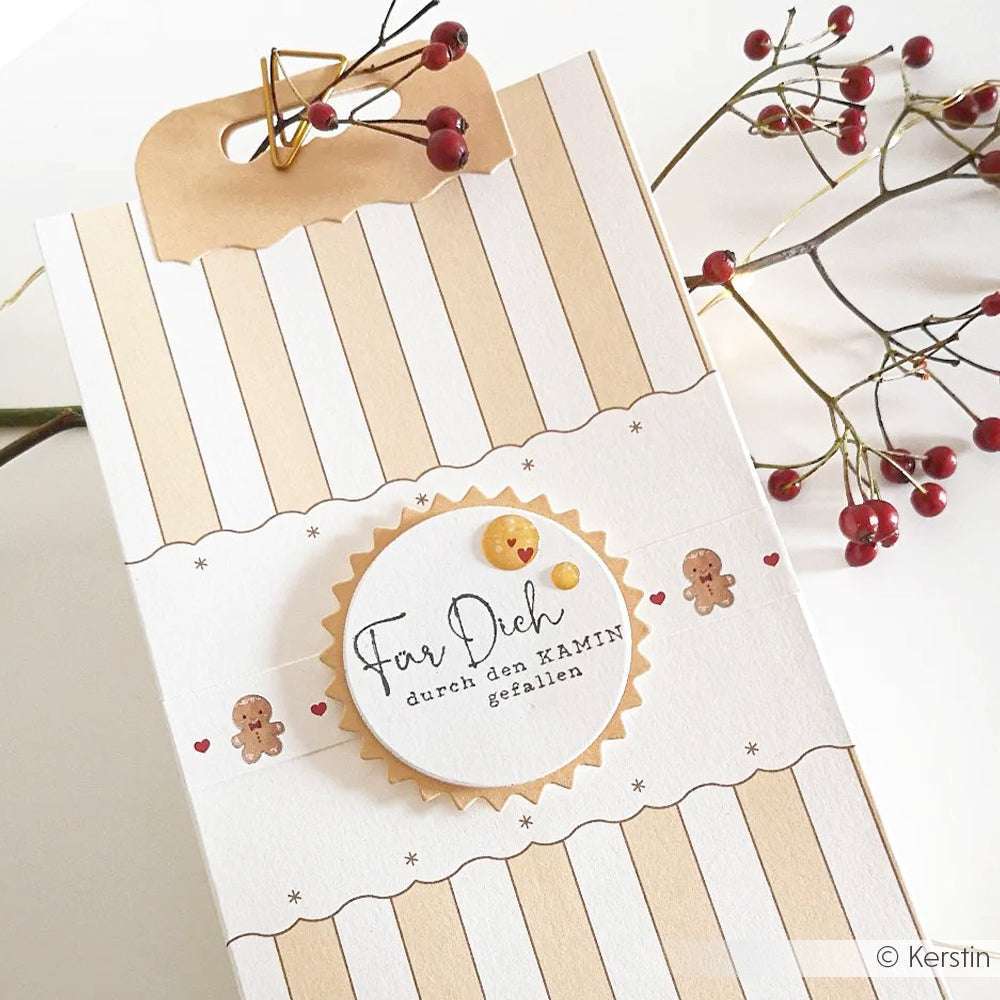 Gestreifte Geschenkschachtel aus Designpapier von Alexandra Renke mit deutschem Text, Lebkuchenmännern und roten Beeren auf Zweigen daneben.