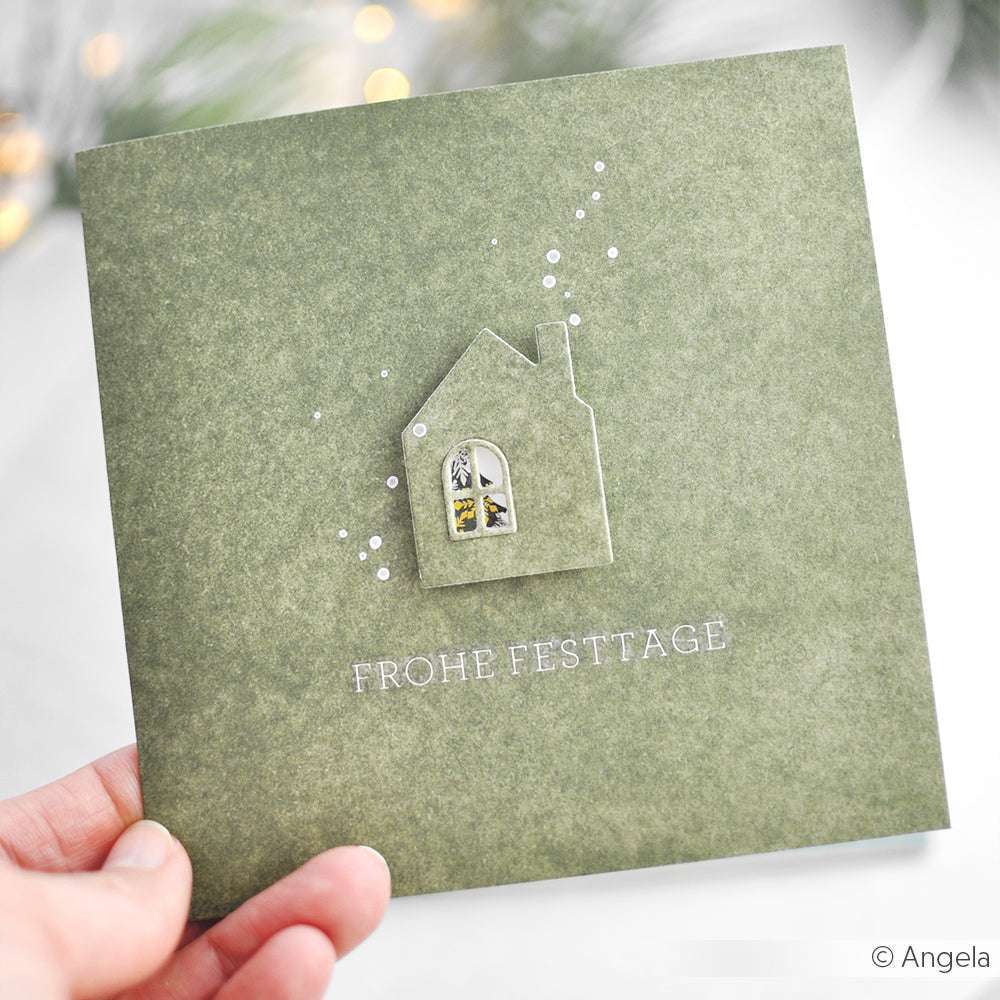 In der Hand hält sie eine grüne Karte von Alexandra Renke Designpapier Mimi Blassgrün, die ein Hausmotiv und Frohe Festtage" zeigt - ein schöner Akzent für festliche Papierbastel- oder Buchbindeprojekte.