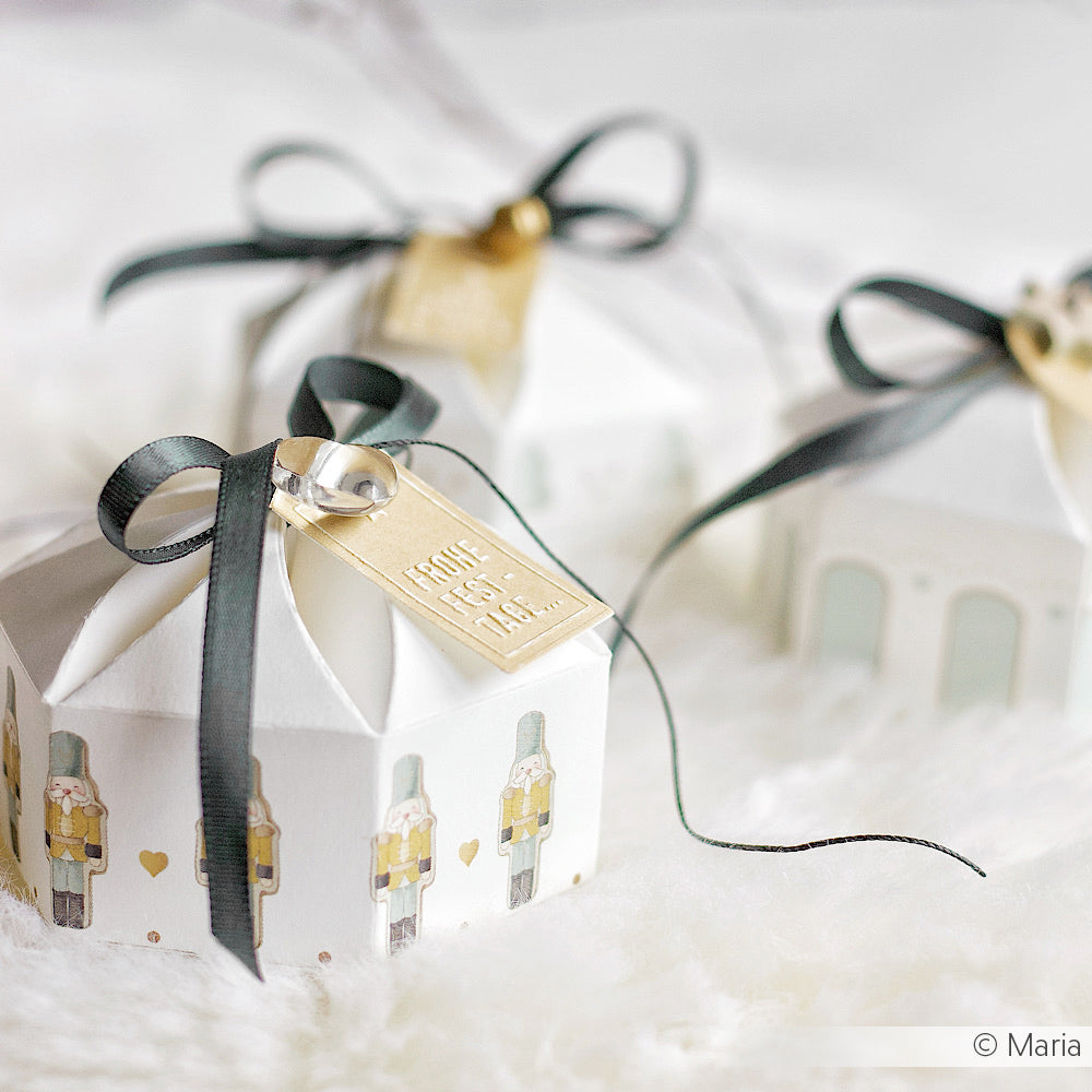 Drei weiße Alexandra Renke Geschenkboxen mit Nussknacker-Motiven auf Designpapier, verziert mit schwarzen Bändern und goldenen Anhängern, ruhen auf einer weichen weißen Oberfläche.