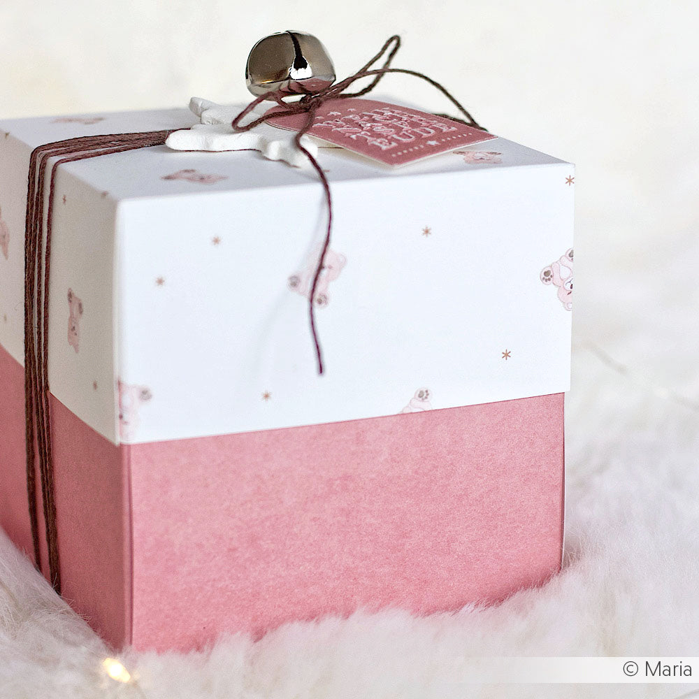 Eine rosa-weiße Geschenkbox mit dem Designpapier Mimi Antikrosa von Alexandra Renke, verziert mit einer Schleife aus Garn und einer Glocke - ideal für Papierprojekte oder Buchbindereien - auf einer weichen, weißen Oberfläche.