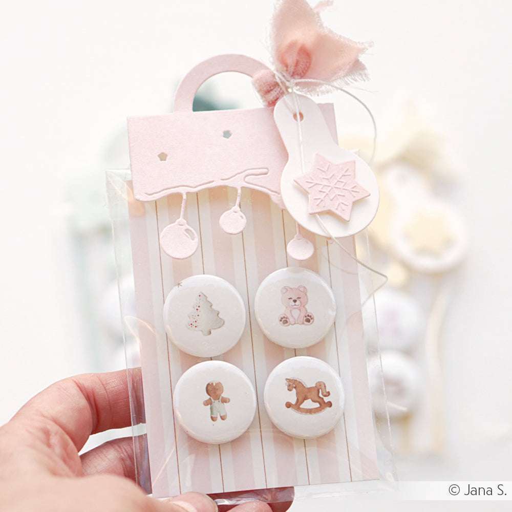 Eine Hand hält eine pastellrosa Geschenktüte von Alexandra Renke, die mit einer Rosa Streifen Bordüre verziert ist und vier dekorierte weiße Kekse mit süßen Weihnachtsmotiven enthält.