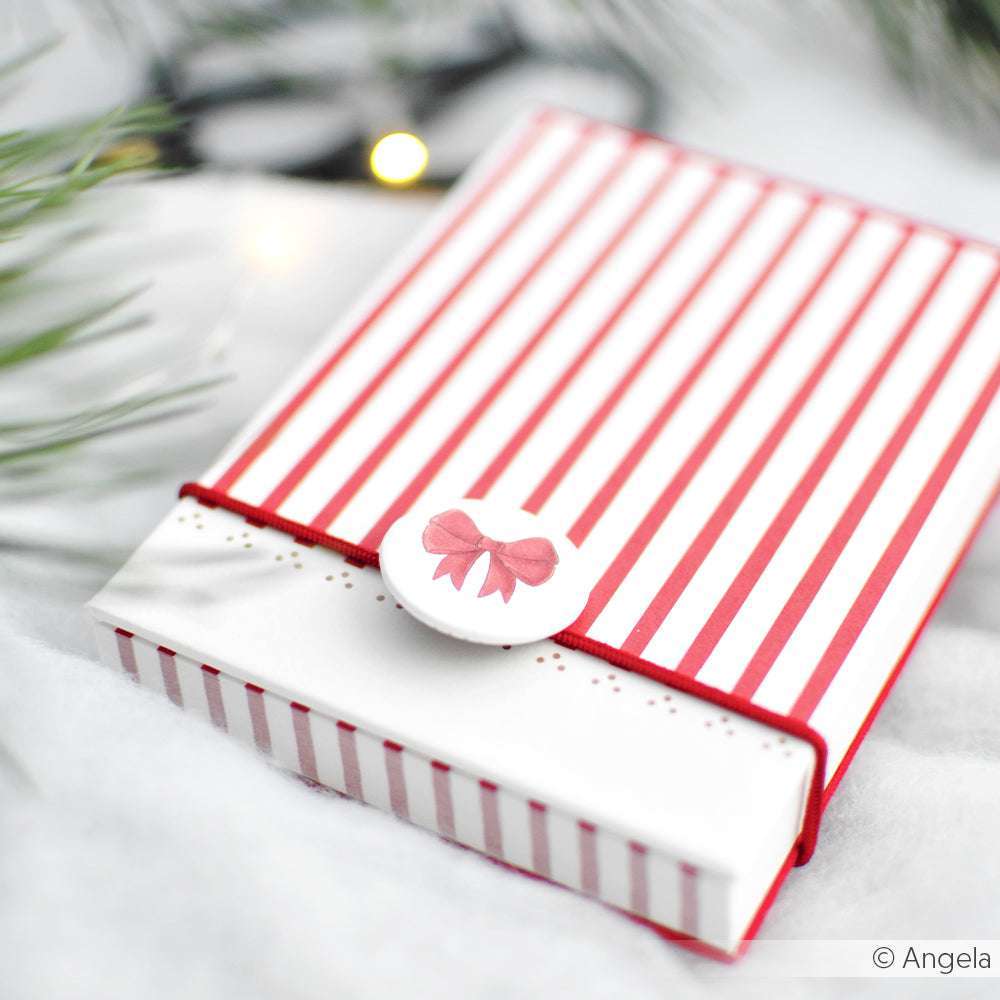 Eine rot-weiß gestreifte Geschenkbox mit rosafarbenem Schleifenverschluss, gefertigt aus Designpapier von Alexandra Renke, ruht auf einer zarten weißen Oberfläche - ideal für festliche Papierprojekte oder als charmante Aufmerksamkeit vom Weihnachtsmann.