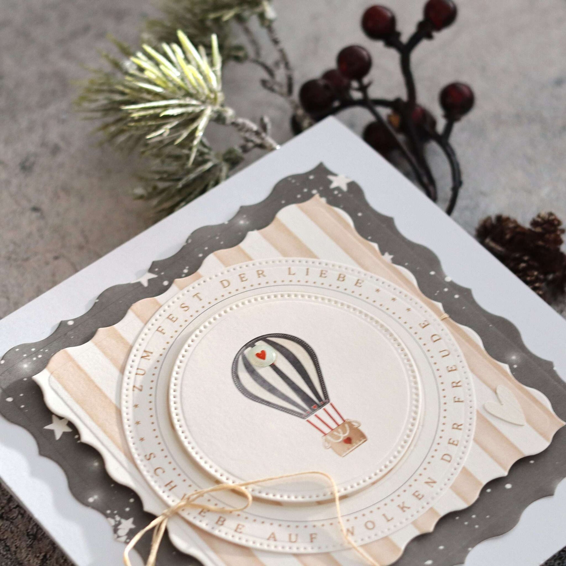 Grußkarte von Alexandra Renke mit einem Heißluftballon-Motiv auf elegantem Papier, mit Schnur gebunden und neben Tannenzweigen und Beeren platziert.