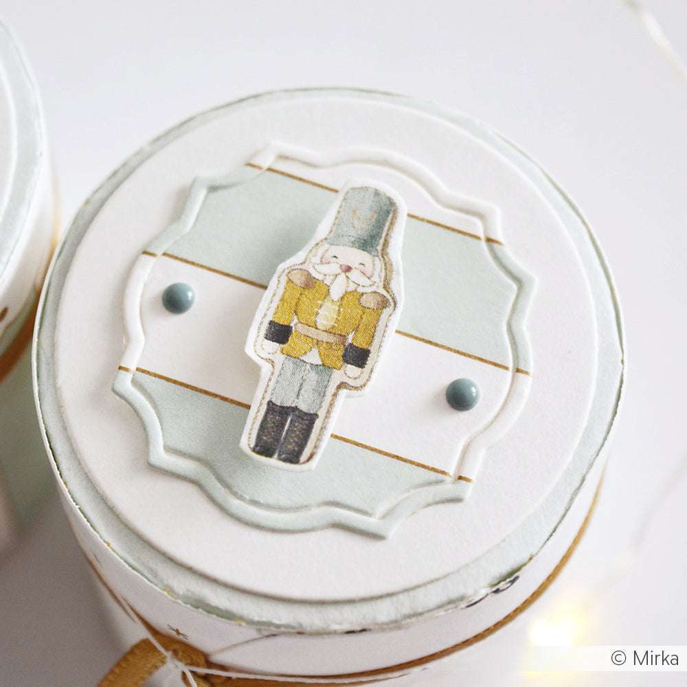 Runder weißer Dosendeckel von Alexandra Renke mit einer dekorativen Nussknacker-Illustration und zwei blauen Punkten - ideal für Papierprojekte oder als Inspiration für Grußkarten mit Designpapier.