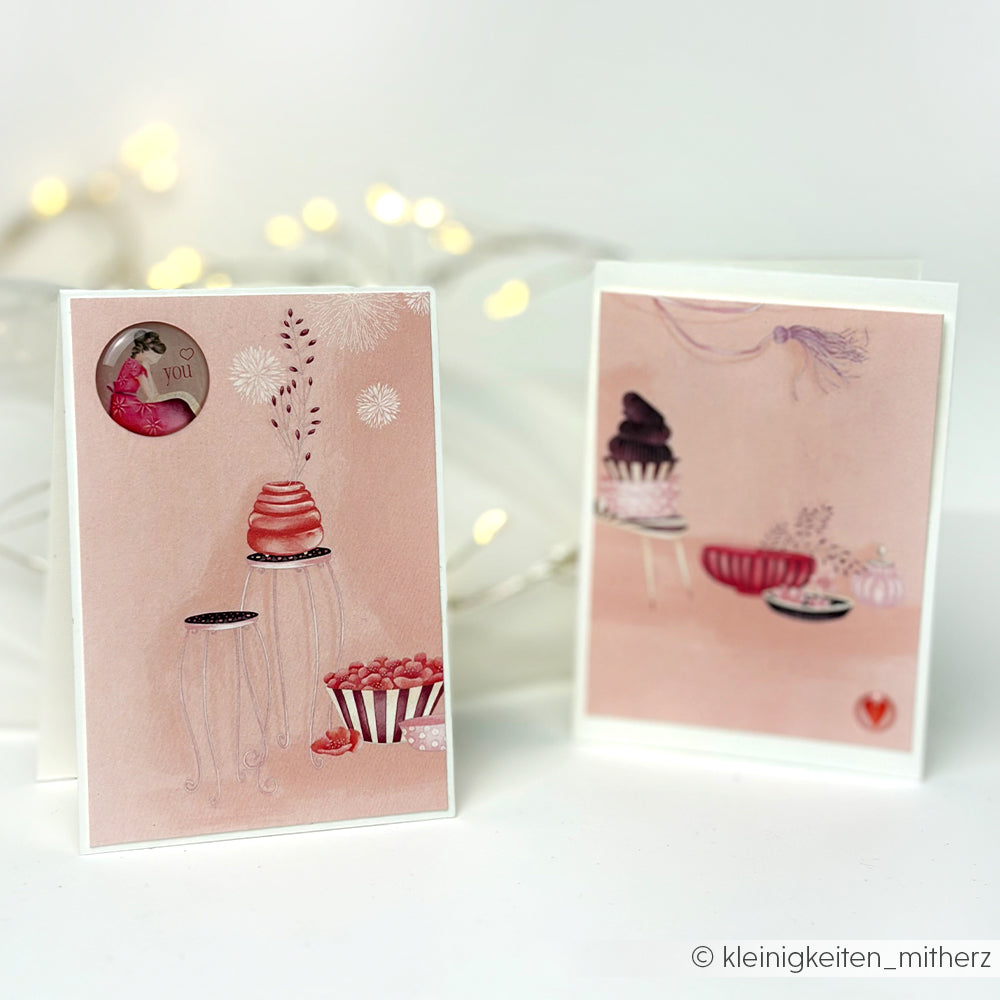 Zwei pastellrosa Alexandra Renke-Grußkarten mit skurrilen Dessert-Illustrationen aus Designpapier stehen auf einer weißen, mit Lichterketten geschmückten Fläche.