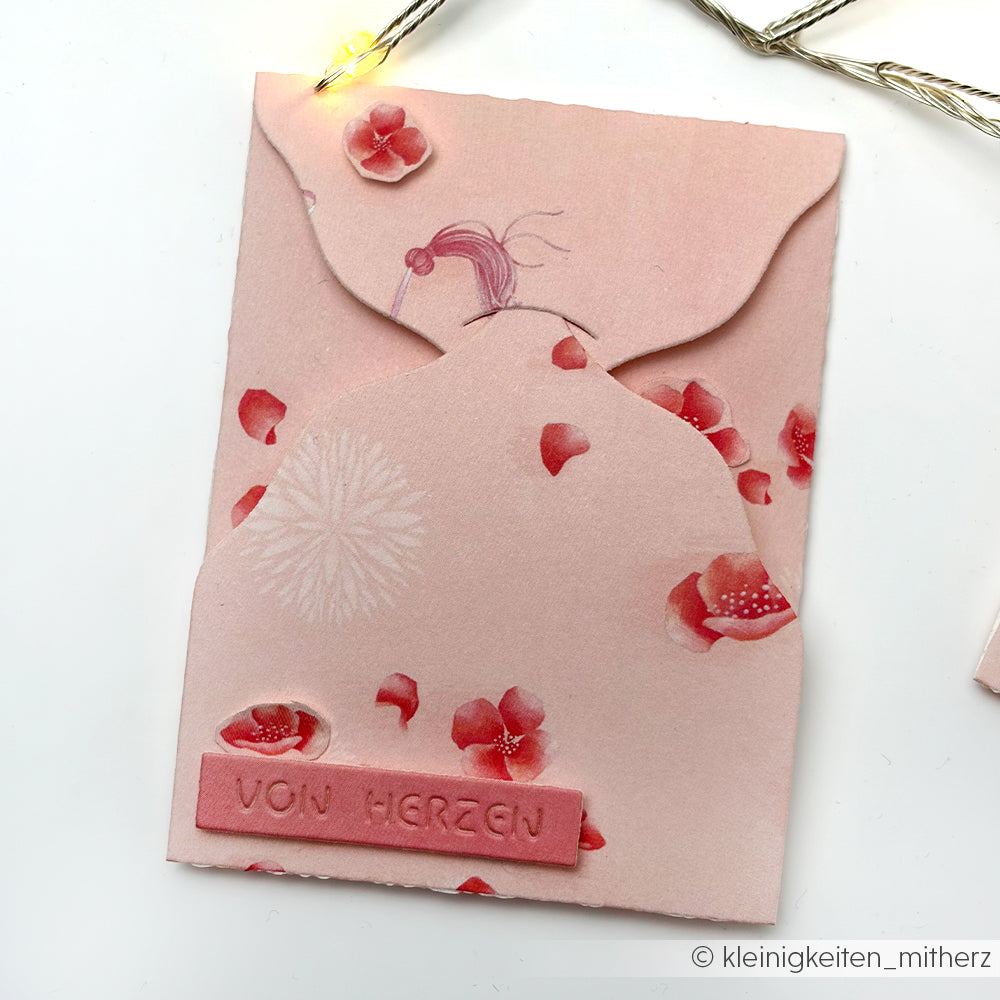 Rosa geblümter Briefumschlag von Alexandra Renke aus Designpapier mit geriffelter Klappe und "VON HERZEN"-Etikett - perfekt für Papierprojekte, die von Lisas Figurinen Blütenzauber inspiriert sind. Abgebildet auf weißem Hintergrund.