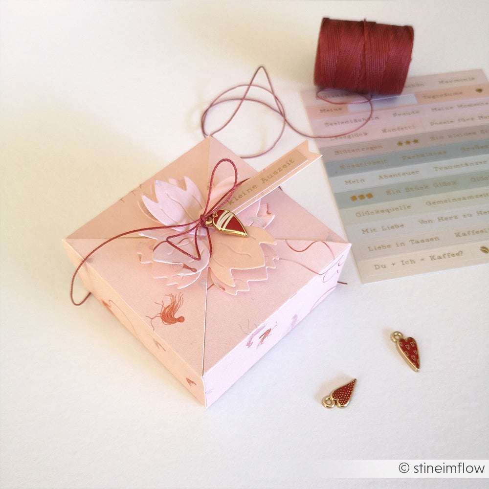 Eine rosafarbene Alexandra Renke-Geschenkbox mit Blumenmuster und Kordel sitzt neben Anhängern, einer Garnrolle, Etikettenaufklebern und elegantem Designpapier - ideal für kreative Papierprojekte und durchdachte Geschenkideen.