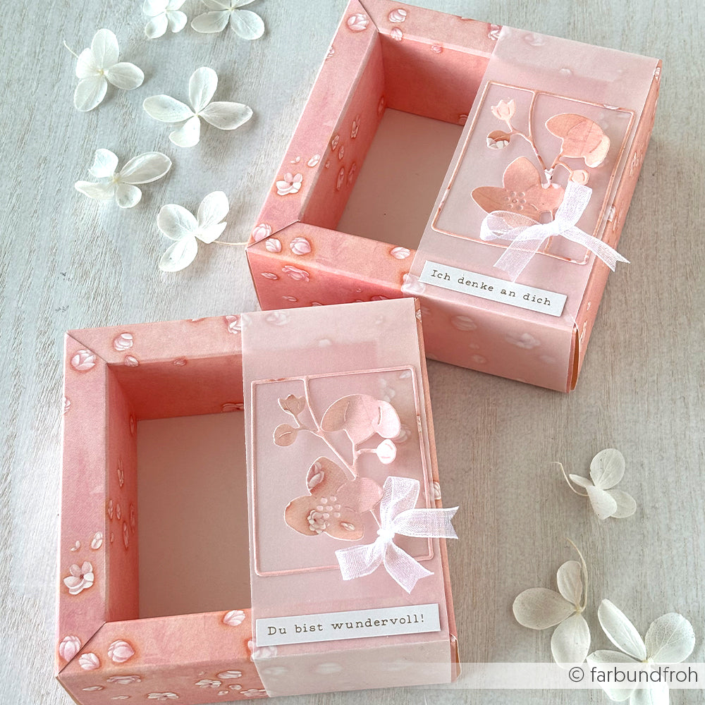 Zwei rosafarbene, mit Alexandra Renke Designpapier gestaltete Geschenkboxen mit deutschen Botschaften, Schleifenband und weißen Zierblumen in der Nähe.