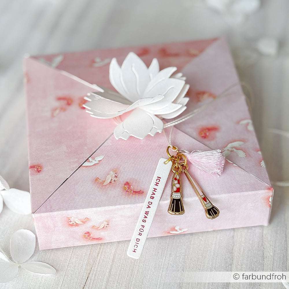 Eine rosafarbene, geblümte Geschenkschachtel von Alexandra Renke, hergestellt aus Designpapier und verziert mit einer weißen Papierblume, Anhängern und einer Quaste - ideal für Papierprojekte oder zur Präsentation von Lisas Figurinen Vogelschwarm.