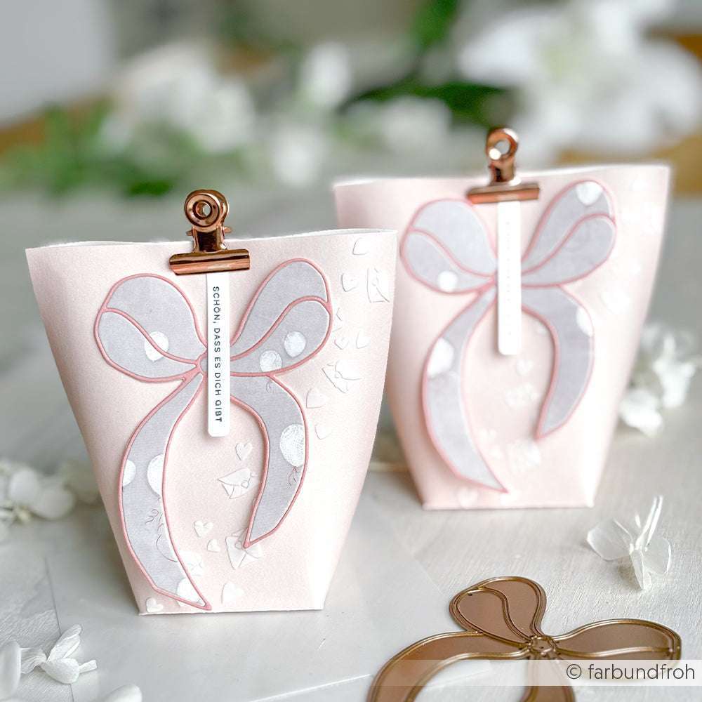 Zwei pastellrosa Geschenktüten von Alexandra Renke mit großen Schleifenmotiven, gefertigt aus elegantem Designpapier und mit goldenen Clips befestigt, auf weißem Untergrund mit Blütenblättern.