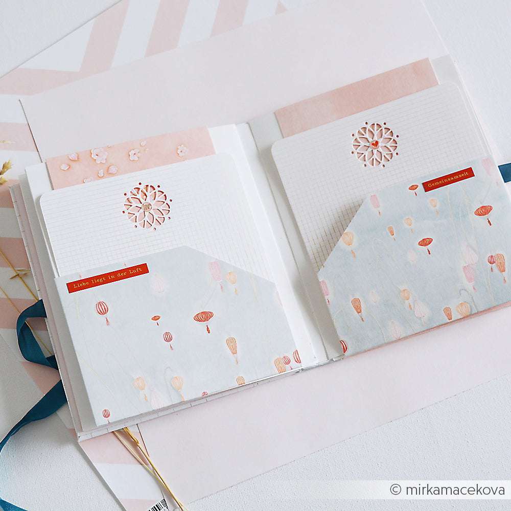Alexandra Renke offenes Scrapbook mit gemusterten Taschen, Notizkarten und pastellfarbenem Designpapier - ideal für Papierprojekte und kreative Buchbindereien - auf einer weißen Fläche.