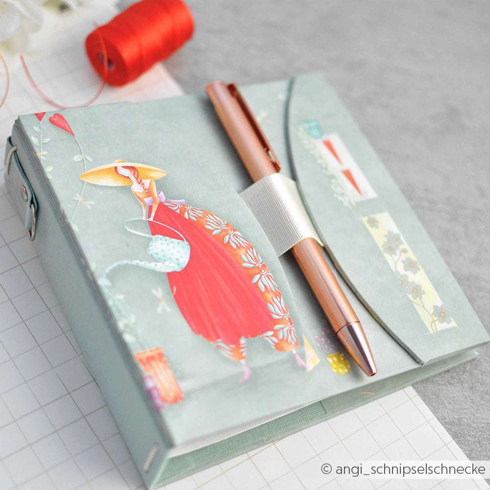 Ein Stift ruht auf einem pastellfarbenen Notizbuch von Alexandra Renke Designpapier, das Lisas Figurinen Gartenzauber zeigt - eine Frau in einem roten Kleid; eine Fadenspule im Hintergrund verleiht Papierprojekten zusätzlichen Charme.