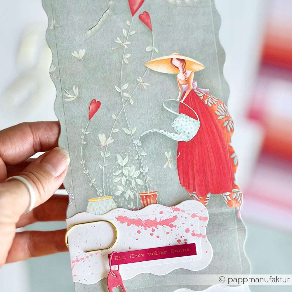 In der Hand hält er eine Alexandra Renke Grußkarte mit einer Frau in Rot und Herzen, auf der "Ein Herz voller Sonne" zu lesen ist - die perfekte Inspiration für Designpapier oder Papierprojekte.