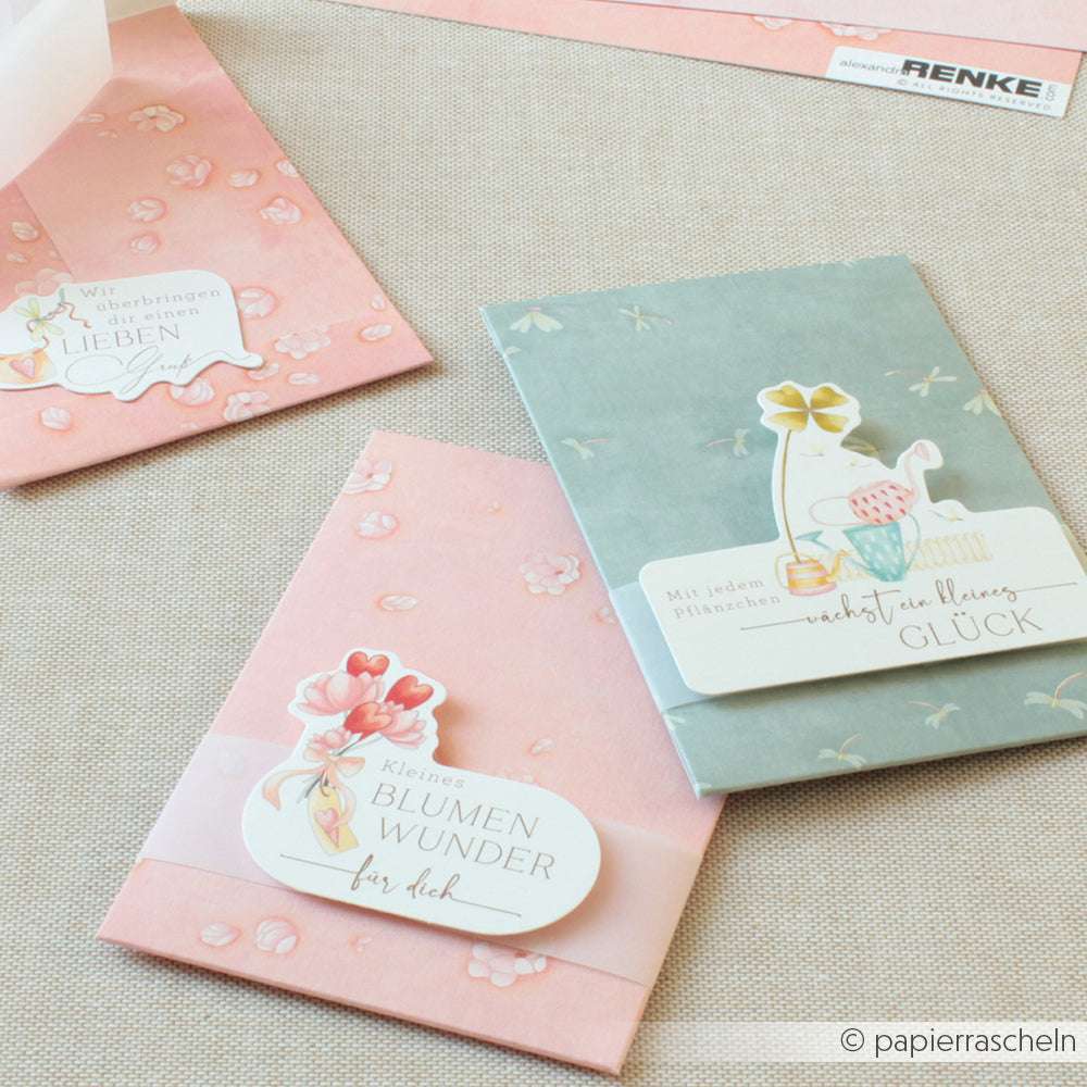 Drei pastellfarbene florale Briefumschläge mit illustrierten Karten und deutschem Text von Alexandra Renke, gefertigt aus hochwertigem Designpapier, gestaltet auf einer leichten Stoffoberfläche.