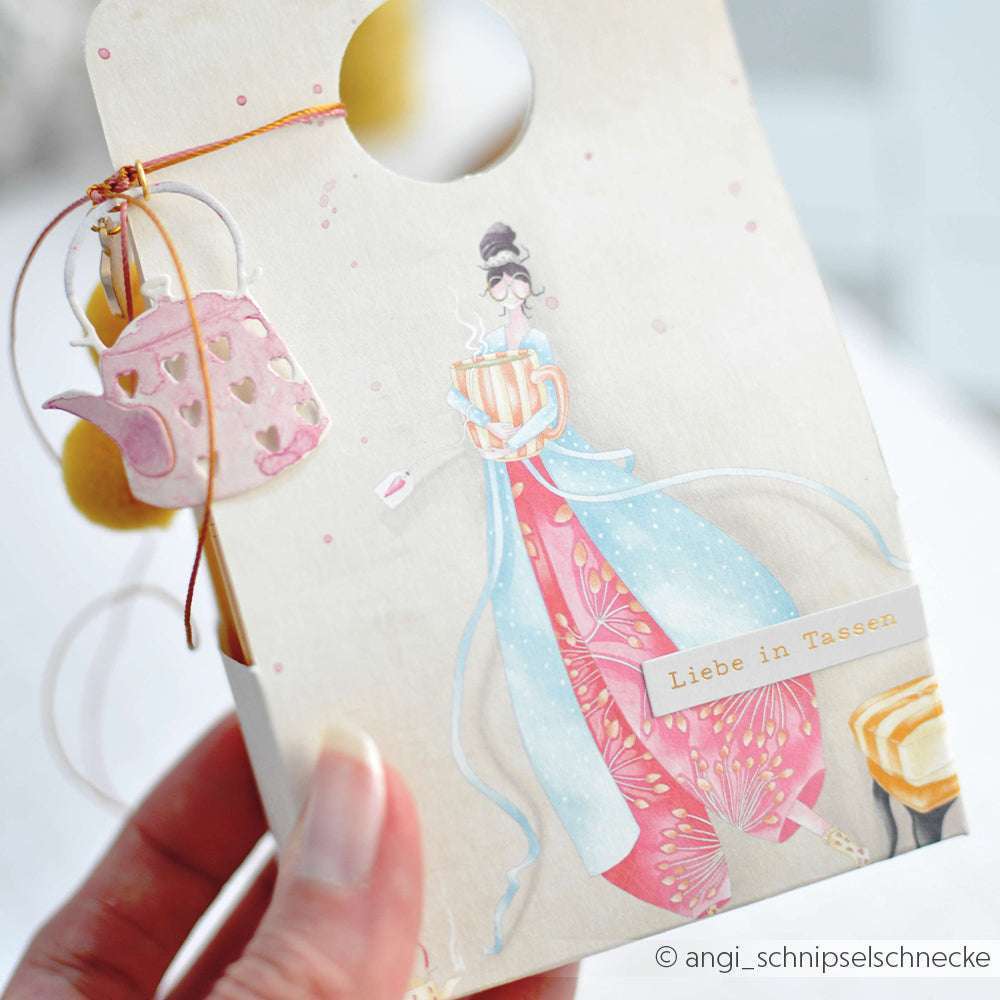 Eine Hand hält eine mit Alexandra Renke Designpapier gestaltete Karte, die eine Dame in einem rosa Kleid, einen Teekannenanhänger und den Text "Liebe in Tassen" zeigt.