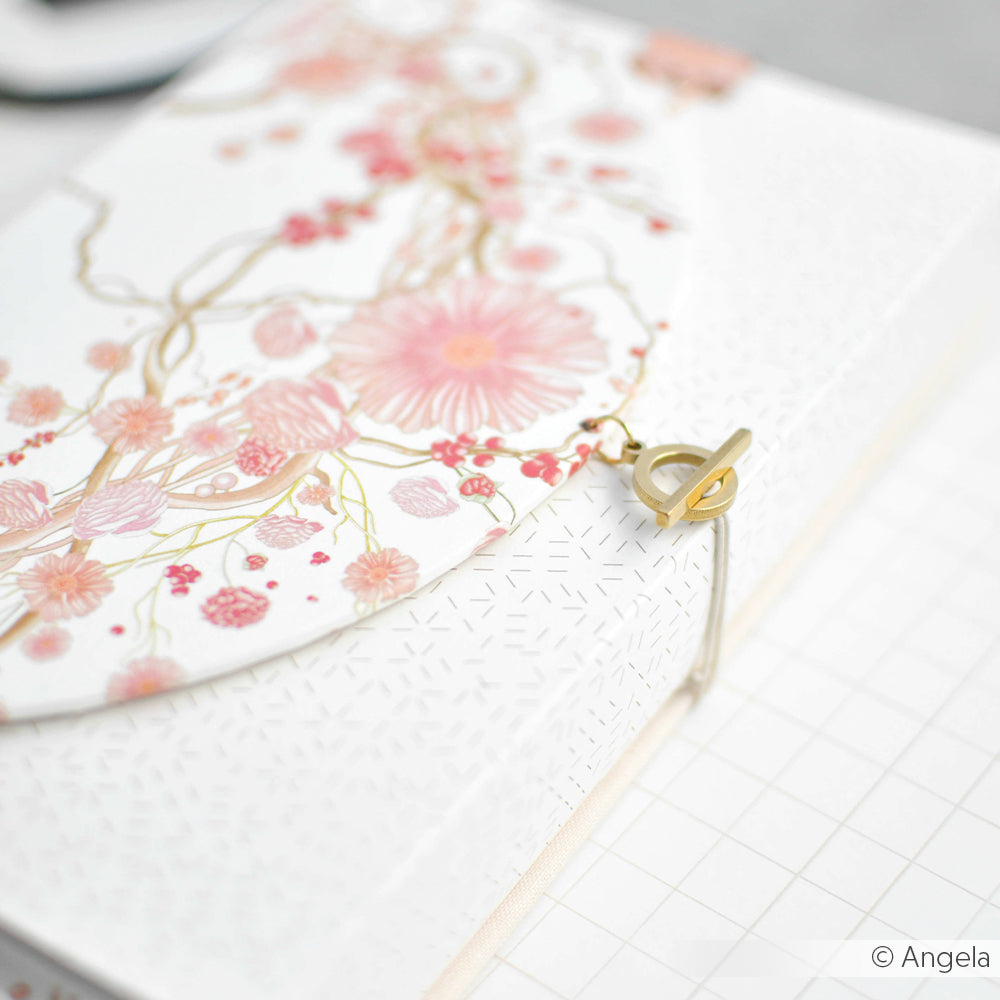 Das weiße Notizbuch von Alexandra Renke mit rosafarbenem Blumenmuster und goldener Schließe aus feinem Designpapier ruht auf einer gitterförmigen Oberfläche - eine elegante Wahl für Ihre Papierprojekte.