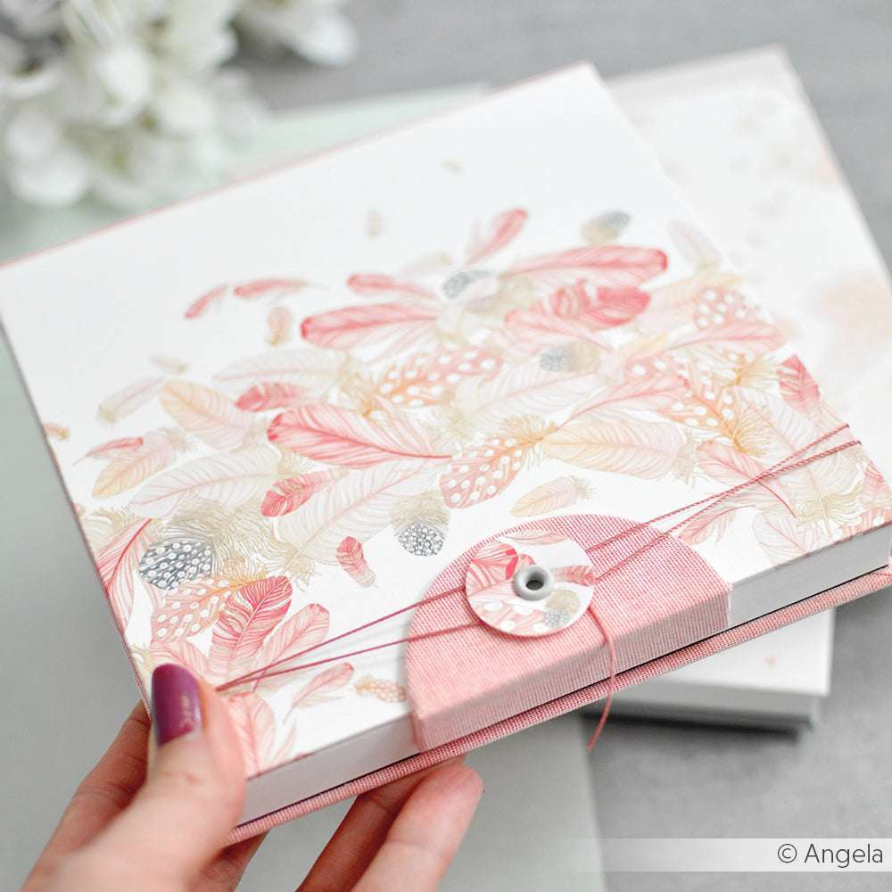 Eine Hand hält einen verzierten Umschlag aus elegantem Alexandra Renke Designpapier mit rosa und goldenen Federmotiven und einem Kordelverschluss.