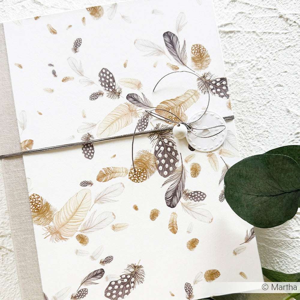 Eine Karte aus Alexandra Renkes Designpapier Flauschiger Federwirbel, akzentuiert mit Schnur und einer weißen Perle, liegt neben grünen Blättern auf einer strukturierten Oberfläche - ideal für Buchbinderei oder Papierprojekte.