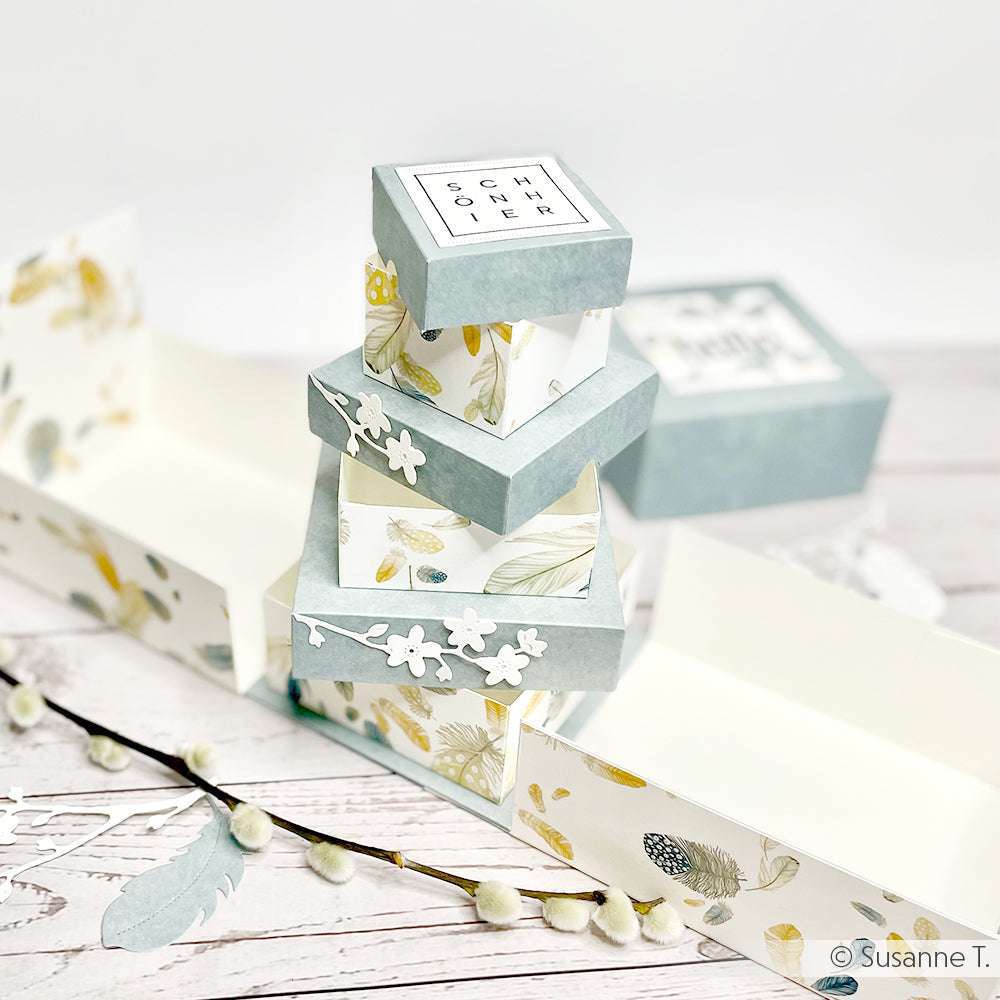 Gestapelte pastellfarbene Geschenkschachteln mit floralen Motiven von Alexandra Renke aus hochwertigem Designpapier, umgeben von offenen Schachteln und dekorativen Zweigen auf einer weißen Fläche.