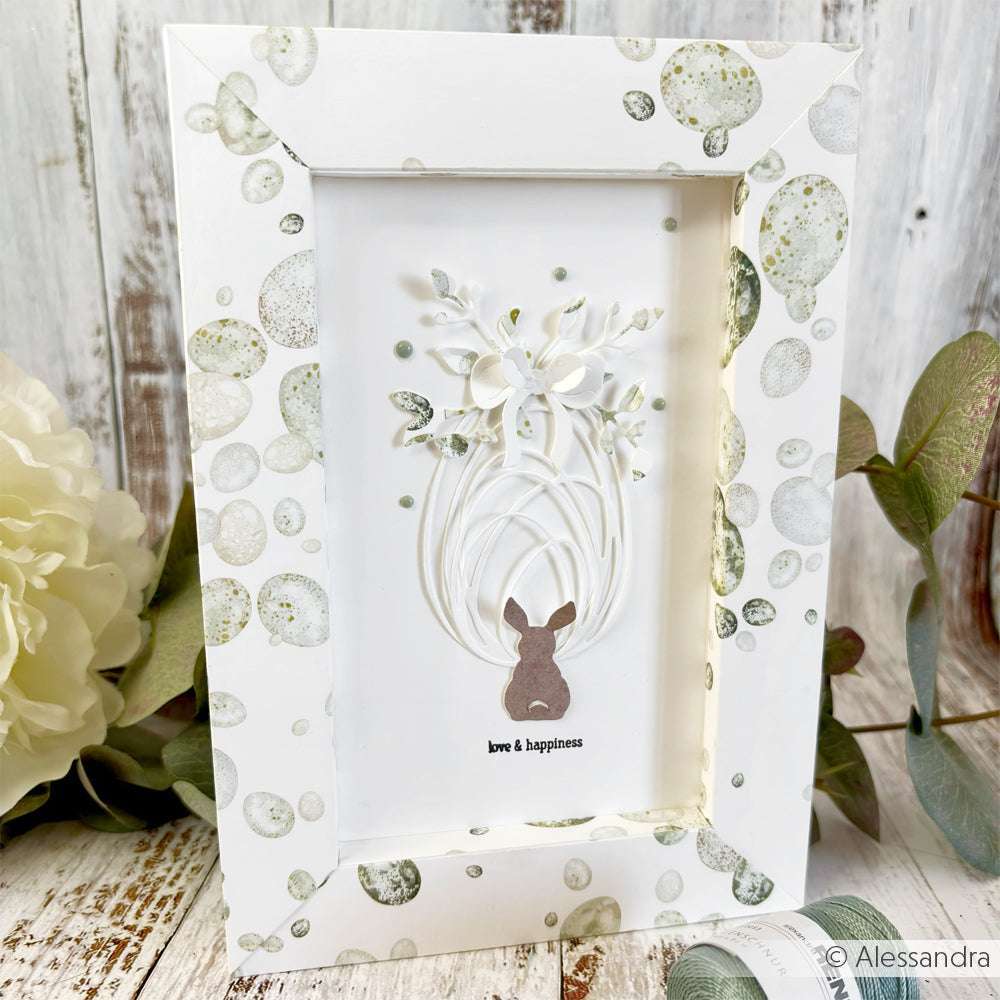 Gerahmte Kunst von Alexandra Renke: eine Häschensilhouette unter weißen Papierblumen mit dem Text "Liebe & Glück" auf gepunktetem Designpapier - ideal für Grußkarten oder kreative Papierprojekte.