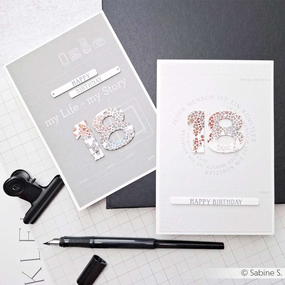 Zwei handgefertigte Happy-Birthday-Karten mit der Nummer 18, hergestellt mit Alexandra Renke Designpapier TypoArt My Story, ausgestellt mit einem Stift auf einem Schreibtisch mit Gittermuster.