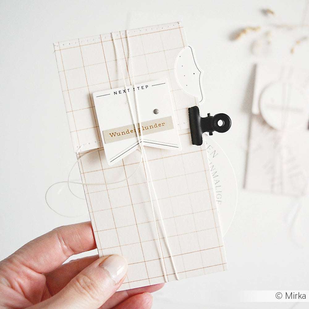 Eine Hand hält eine minimale, gittergemusterte Karte aus Alexandra Renkes Bleistiftkaro Gold Designpapier mit weißen und goldenen Akzenten, die auf der Vorderseite befestigt sind - ideal für Ihre nächsten Papierprojekte.