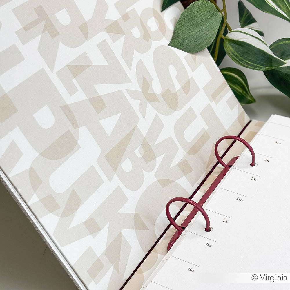 Offener Ringordner von Alexandra Renke mit nach Tagen beschrifteten Trennblättern aus Designpapier, die neben einer grünen Blattpflanze auf einer weißen Fläche stehen.