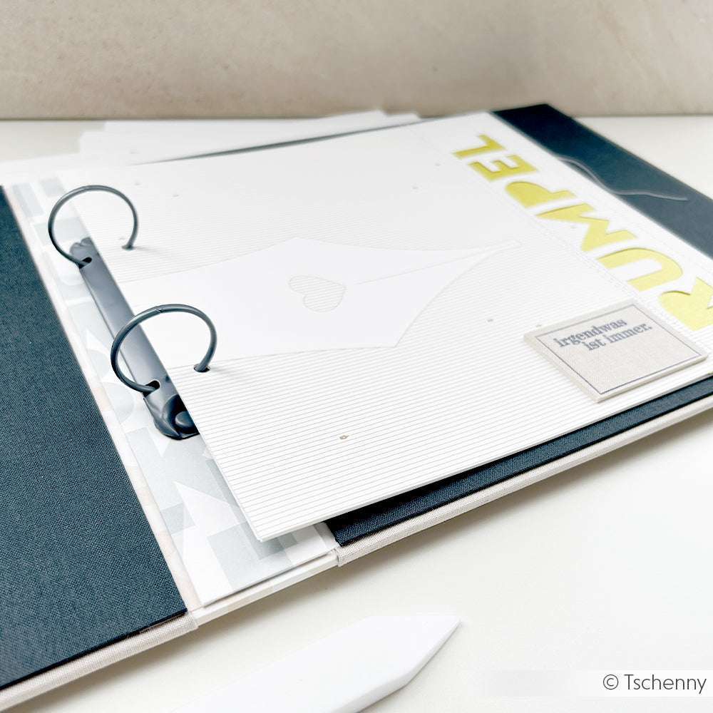 Offenes Scrapbook von Alexandra Renke mit Ringbuch, weißen Seiten und teilweise sichtbarem gelben "RUMPEL"-Text - ideal für Papierprojekte oder passend mit Federspitze auf Liniert Gold Designpapier.