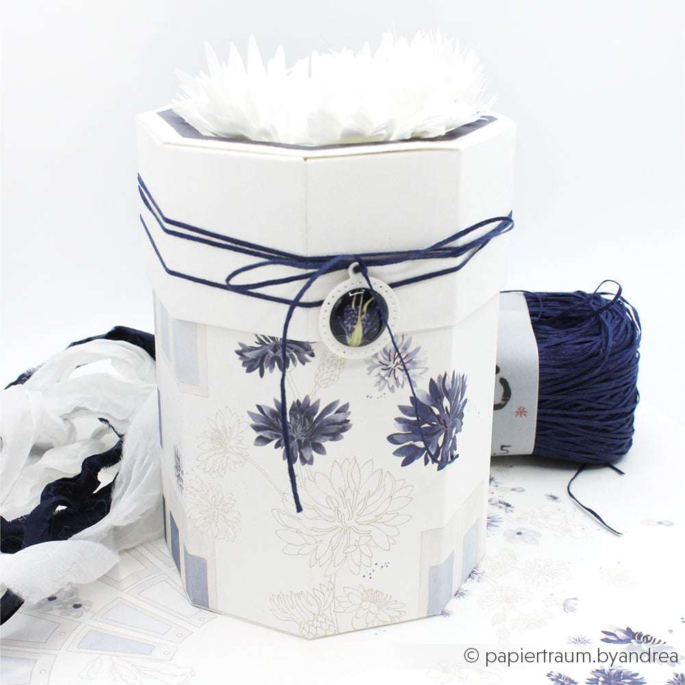 Sechseckiger weißer Geschenkkarton von Alexandra Renke mit floralen Motiven, blauer Kordel, einer Blüte oben und Designpapier-Details - perfekt für Papierprojekte. Mit Farbkartenkreis Blau als Akzent und dunkelblauem Garn daneben.