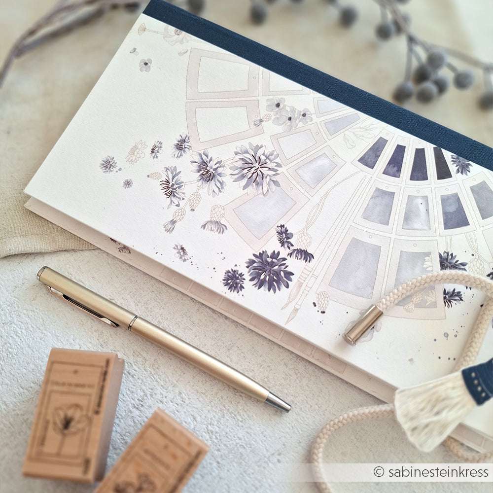 Elegantes florales Notizbuch, silberner Stift und Stempel von Alexandra Renke auf einer hellen Oberfläche mit einer Quaste - eine ideale Inspiration für Papierprojekte oder in Kombination mit dem Farbkartenkreis Blau Designpapier.