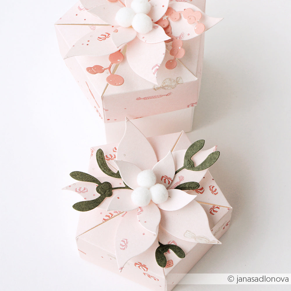 Zwei hellrosa Geschenkboxen für Papierprojekte, verziert mit Weihnachtssternen und weißen Pompons.