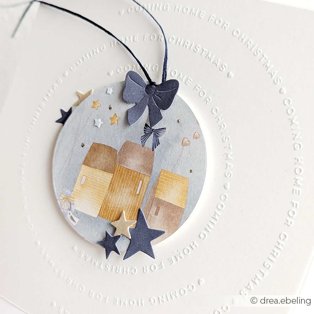 Rundes Ornament mit Häusern, Sternen und einer Schleife auf einer weißen Karte mit der Aufschrift COMING HOME FOR CHRISTMAS in einem Kreis - perfekt für Papierprojekte oder zur Kombination mit Alexandra Renke Designpapier und Weihnachtsfiguren.