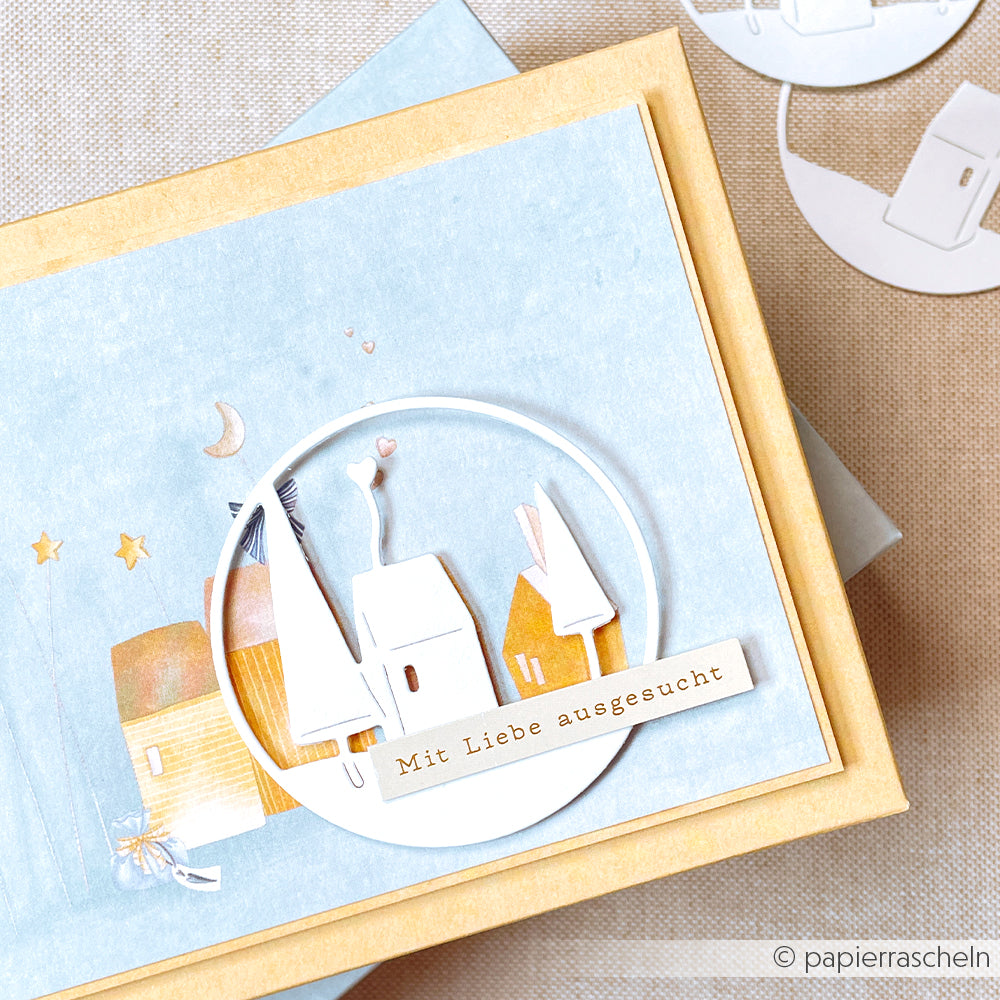 Eine handgefertigte Karte von Alexandra Renke zeigt weiße Papierhäuser und den Schriftzug "Mit Liebe ausgesucht" auf hellblauem Hintergrund - ideal für Papierprojekte oder festliches Flair mit Designpapier Weihnachten.