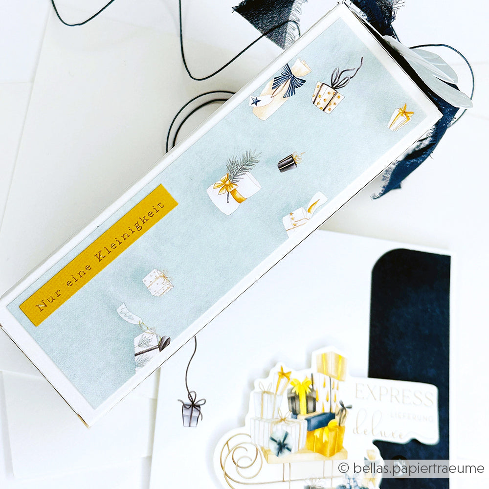 Eine festliche Alexandra Renke Geschenkbox in illustriertem Designpapier mit kleinen Geschenken und einem gelben Etikett mit der Aufschrift "Nur eine Kleinigkeit.