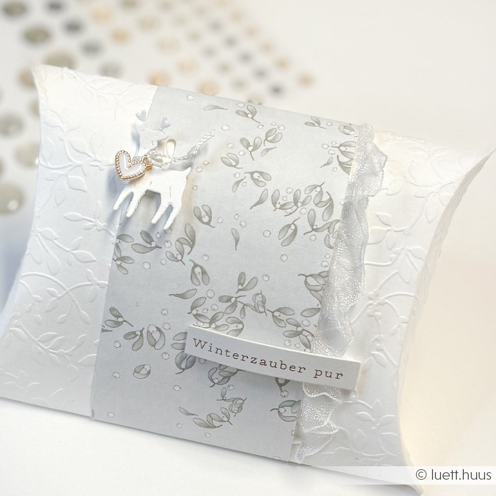 Weiße Geschenkschachtel von Alexandra Renke mit floralem Papier, einem Rentieranhänger, Spitze und einem Schild mit der Aufschrift "Winterzauber pur".