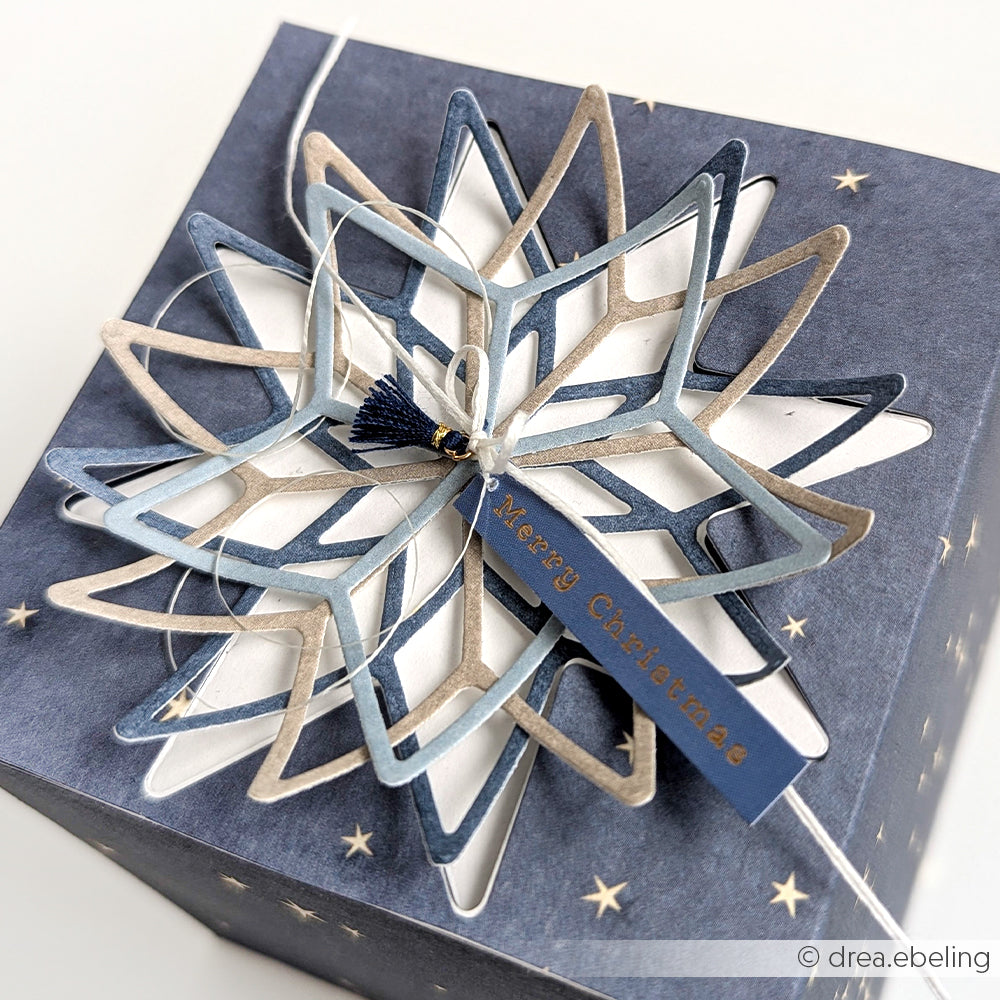 Ein marineblauer Geschenkkarton mit einem geschichteten Alexandra Renke Designpapier Sternenzauber Rauchblau und einem Merry Christmas Anhänger - ideal für festliche Papierprojekte oder Buchbinderei-Kreationen.
